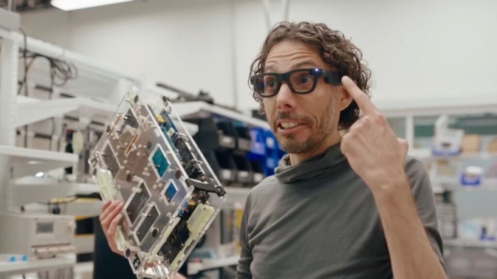 Meta Perkenalkan Aria Gen 2: Kacamata Pintar dengan Sensor Detak Jantung dan Teknologi Canggih 5 Seseorang dengan rambut keriting menunjuk ke kacamata pintarnya dengan satu tangan dan memegang komponen elektronik besar di tangan lainnya. Mereka berdiri di ruang kerja yang penuh teknologi dengan rak dan peralatan di latar belakang.