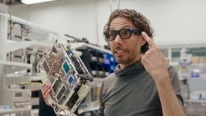 Seseorang dengan rambut keriting menunjuk ke kacamata pintarnya dengan satu tangan dan memegang komponen elektronik besar di tangan lainnya. Mereka berdiri di ruang kerja yang penuh teknologi dengan rak dan peralatan di latar belakang.