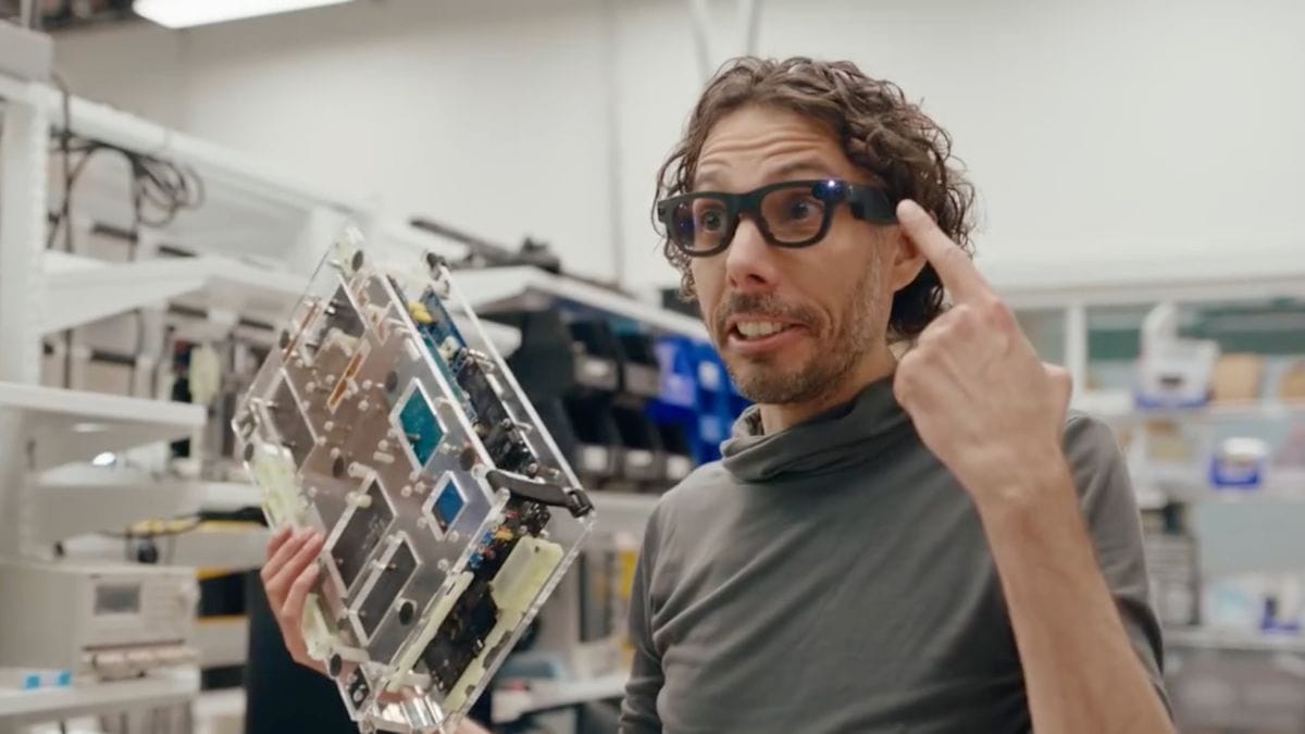 Seseorang dengan rambut keriting menunjuk ke kacamata pintarnya dengan satu tangan dan memegang komponen elektronik besar di tangan lainnya. Mereka berdiri di ruang kerja yang penuh teknologi dengan rak dan peralatan di latar belakang.