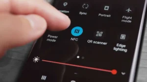Tampilan dekat layar ponsel pintar yang memperlihatkan tangan yang hendak mengetuk ikon fitur NFC. Layar menampilkan ikon-ikon lain, termasuk mode Daya, pemindai QR, dan pencahayaan Edge. Penggeser kecerahan terlihat di bagian bawah.